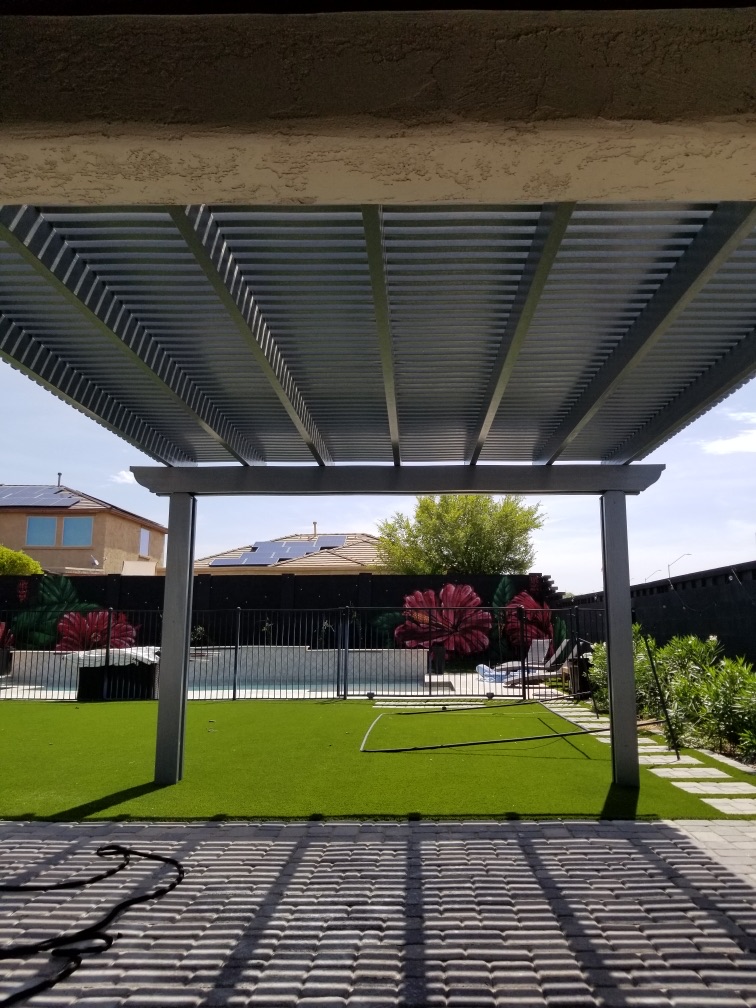 The Meadows (Peoria) under-pergola view of grey alumawood lattice roof over grey paver patio, artificial turf and glass pool fence with hibiscus mural on rear block wall