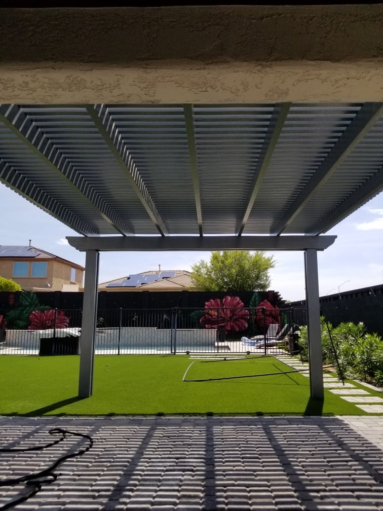 The Meadows (Peoria) under-pergola view of grey alumawood lattice roof over grey paver patio, artificial turf and glass pool fence with hibiscus mural on rear block wall