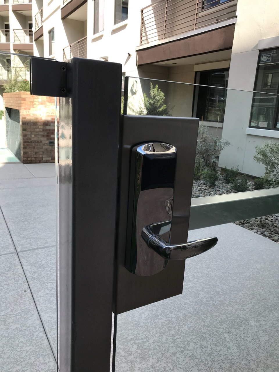 Electronic access reader on a dark-finished gate post beside a glass fence at an apartment pool area.