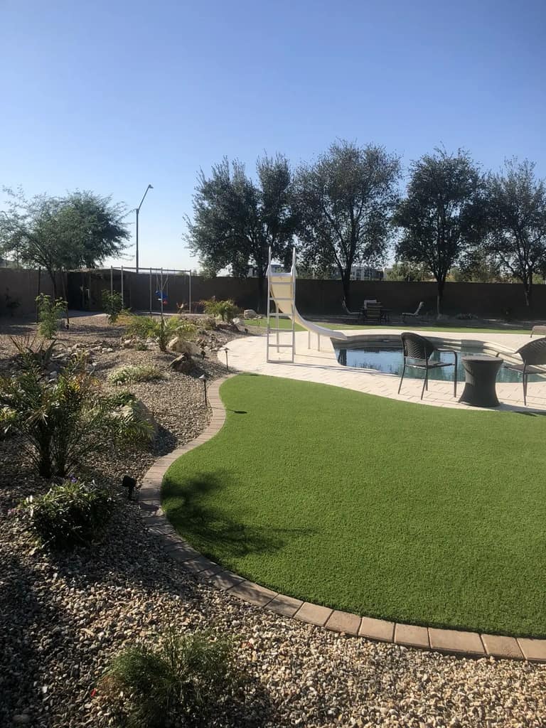 Curved artificial turf beside poolside pavers and a planted desert-rock bed with low-voltage lights.