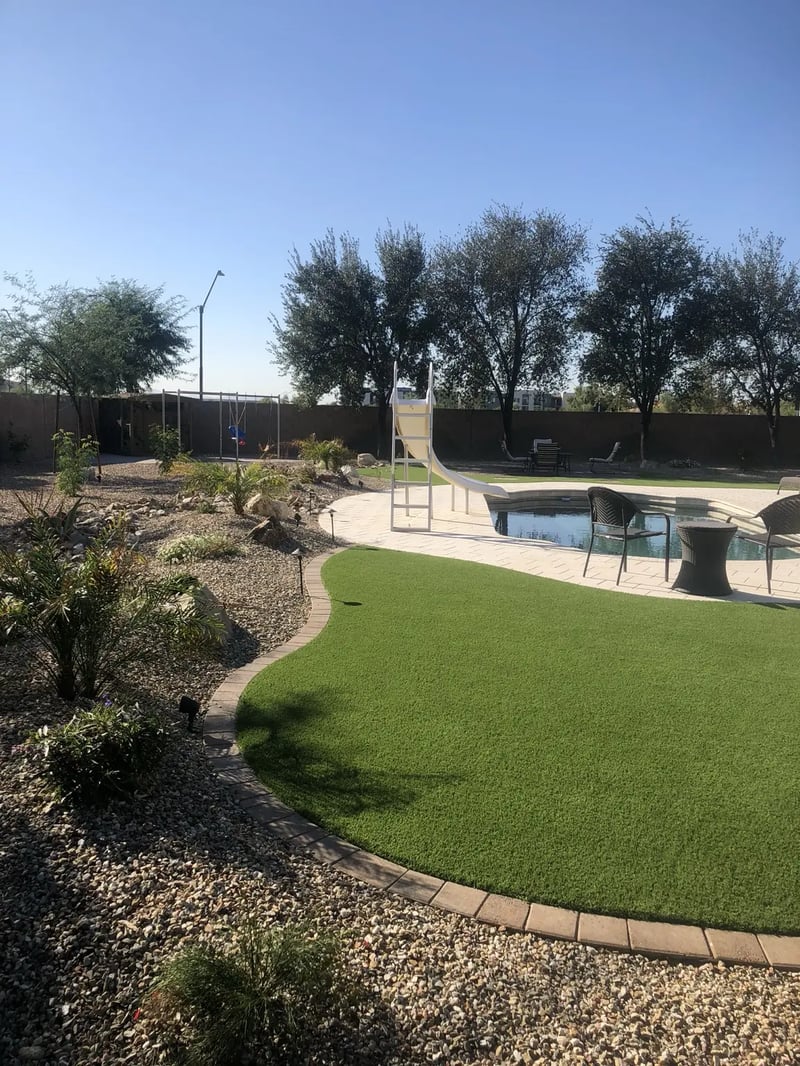 Curved artificial turf beside poolside pavers and a planted desert-rock bed with low-voltage lights.