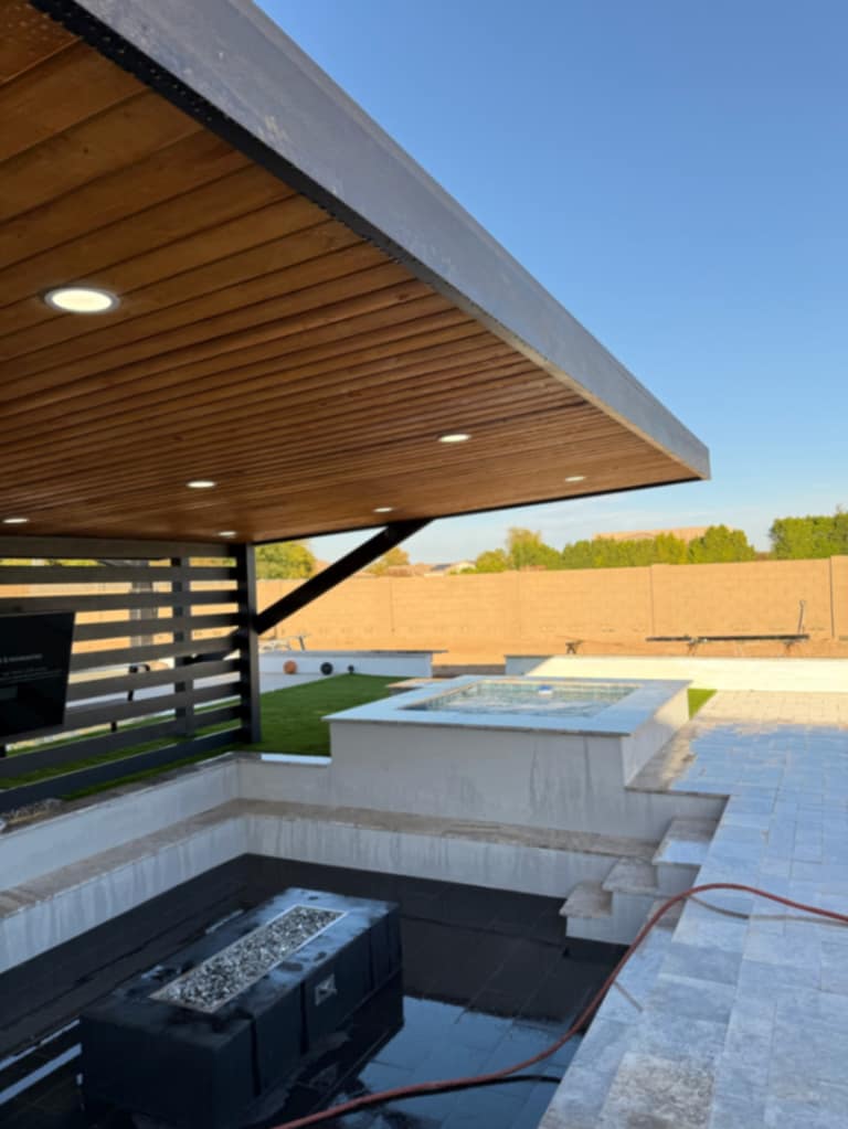 Wide exterior view of black pergola with wood ceiling next to raised spa, sunken fire lounge, and open paver deck.