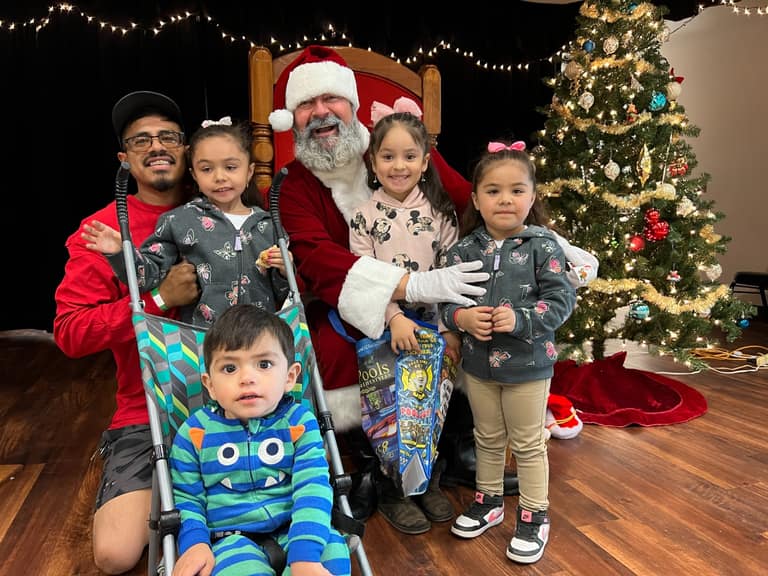 Family of four kids and dad gathered with Santa, holding AE Outdoor Living Luxe Pools and Ugly Pool Guy gift bags at Shop With a Cop