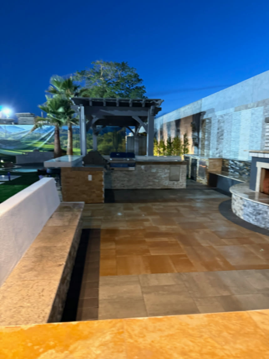 Outdoor kitchen and prep counter beneath pergola beside long illuminated finish wall at dusk
