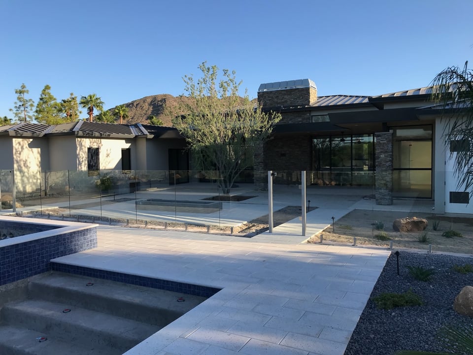 Wide view of a modern estate pool deck bordered by frameless glass fence panels on spigots, beside paver decking with stone architecture behind.
