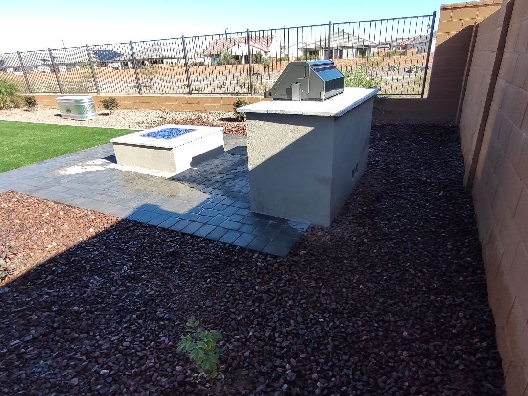 Vistancia (Peoria) side view of stucco outdoor kitchen island and matching square fire pit on charcoal paver patio with artificial turf lawn and wrought-iron view fencing to open desert