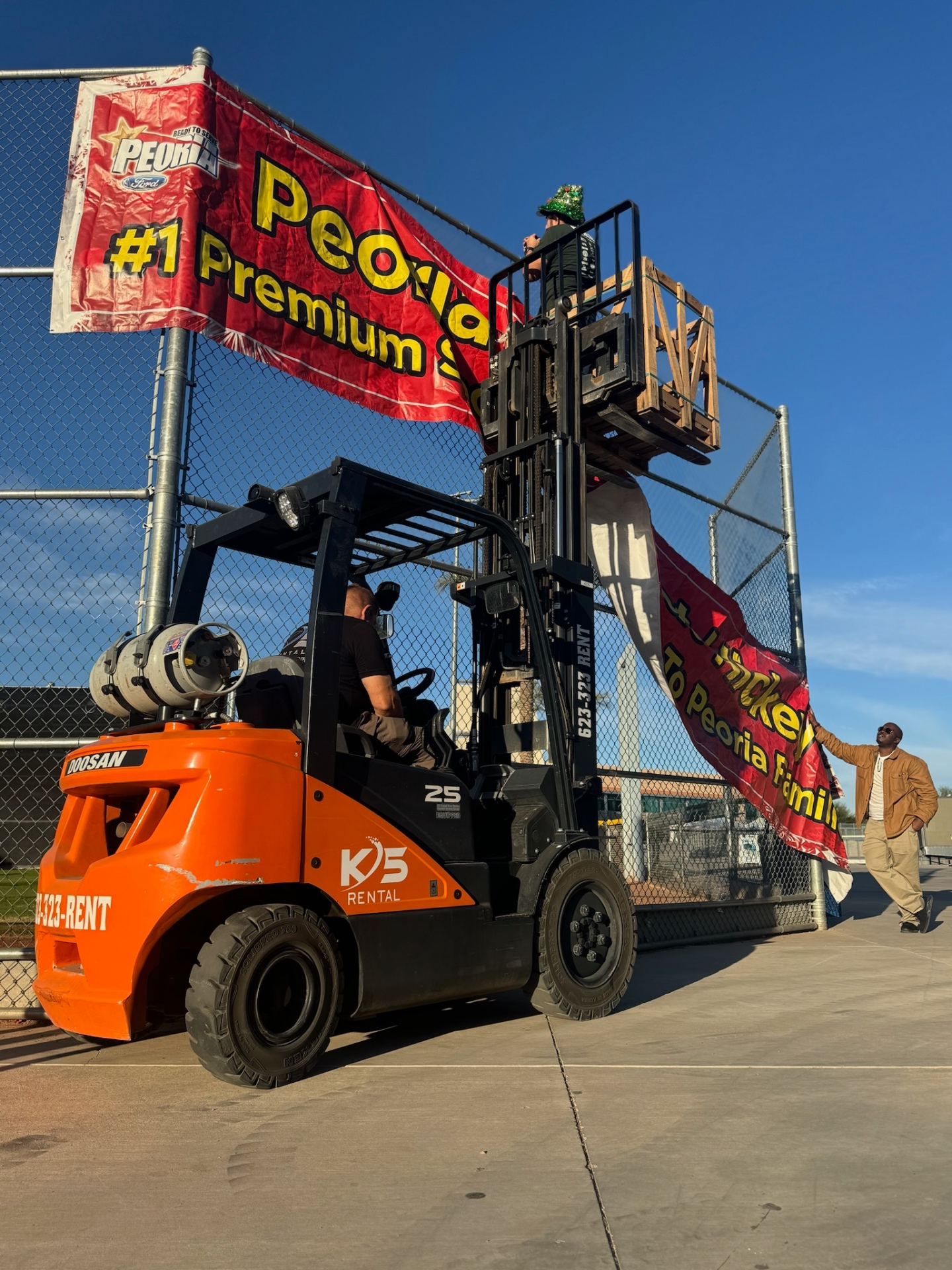 Crew hanging the Day of Giving event banner on the fence at Peoria Sports Complex using a forklift
