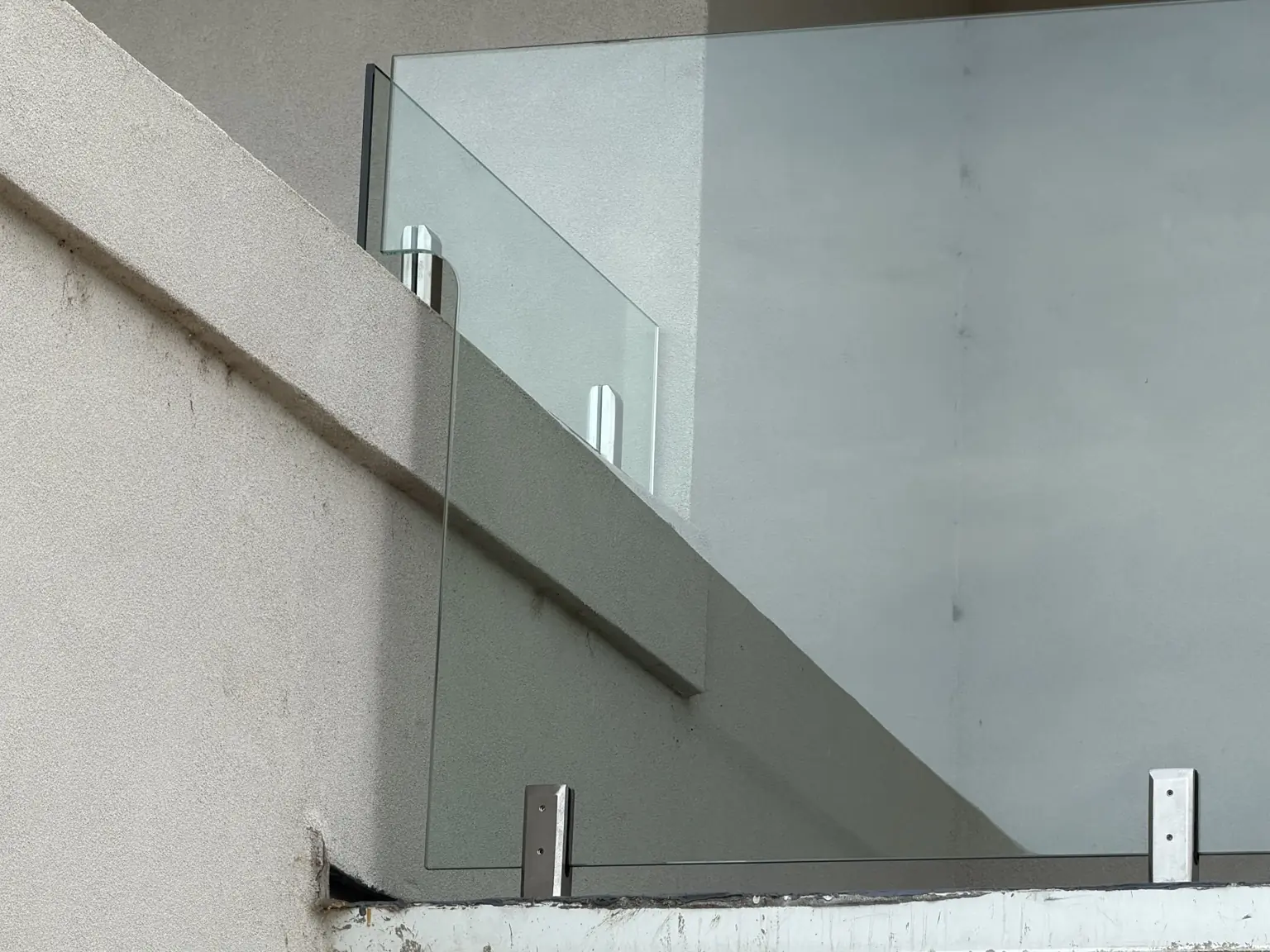 Close-up of glass railing panels held by base-mounted hardware on a stucco balcony.