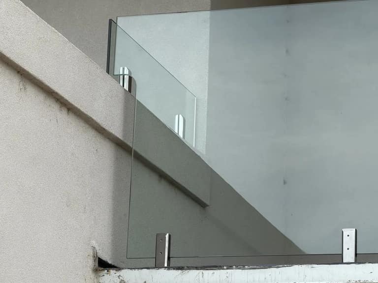 Close-up of glass railing panels held by base-mounted hardware on a stucco balcony.