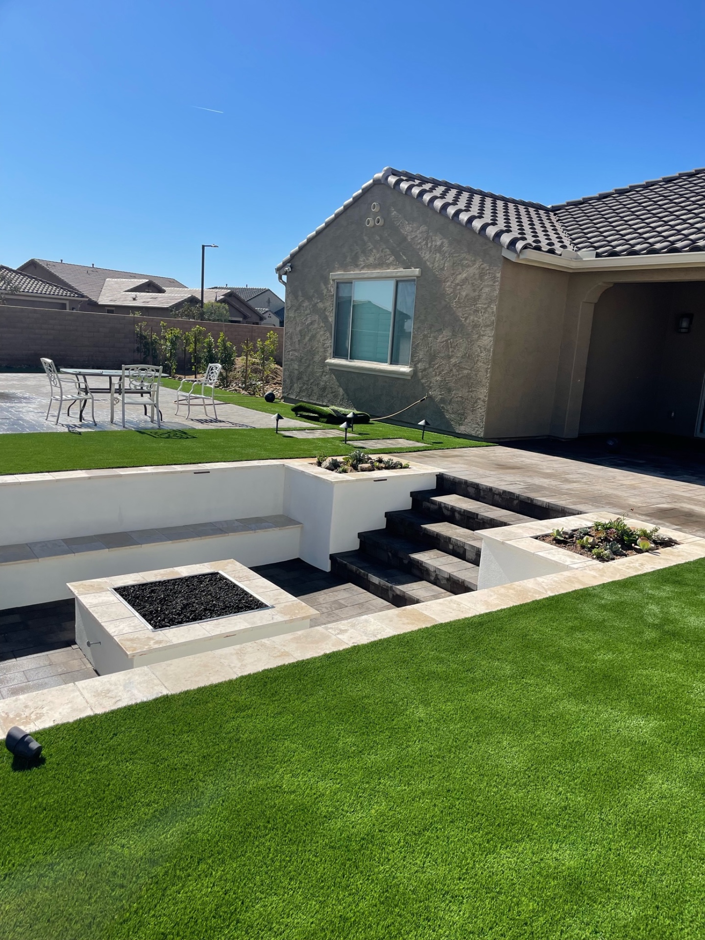Goodyear backyard daytime view of sunken paver firepit lounge with travertine seat walls, planter boxes and lit paver step-down from covered patio