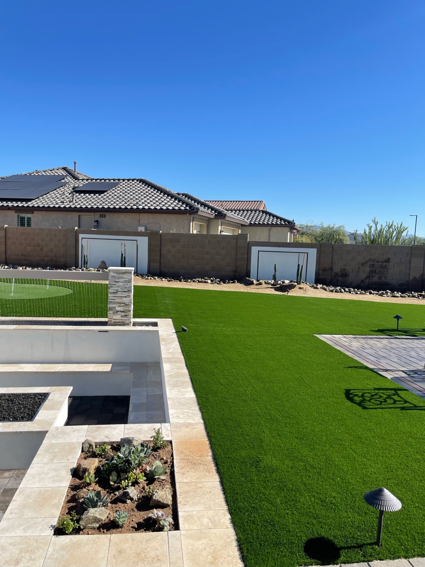 Goodyear backyard artificial turf lawn with putting green, stacked-stone water feature, travertine firepit lounge and paver patio