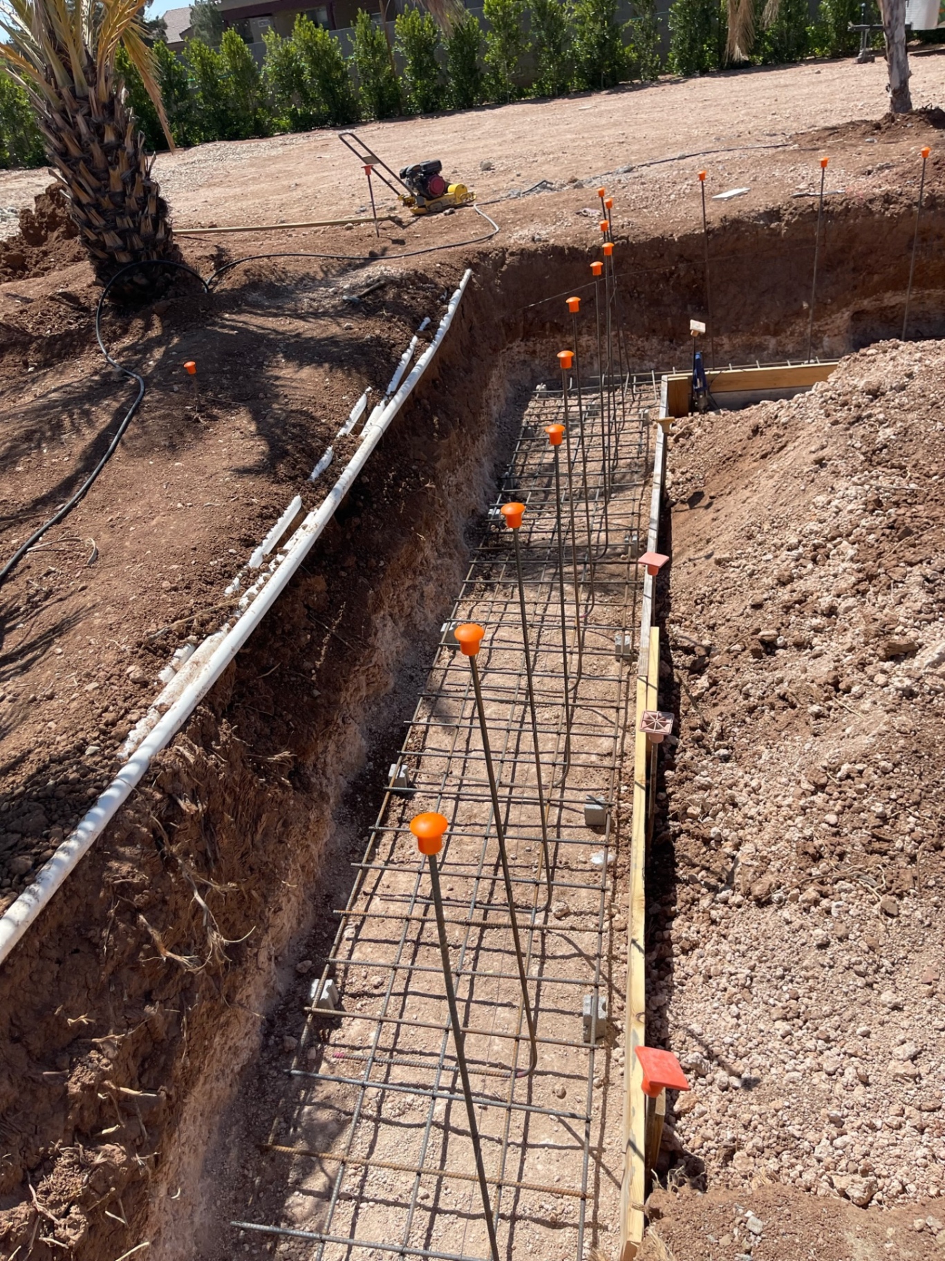 Straight trench footing with a full rebar cage and orange safety caps extending toward the far corner of the yard