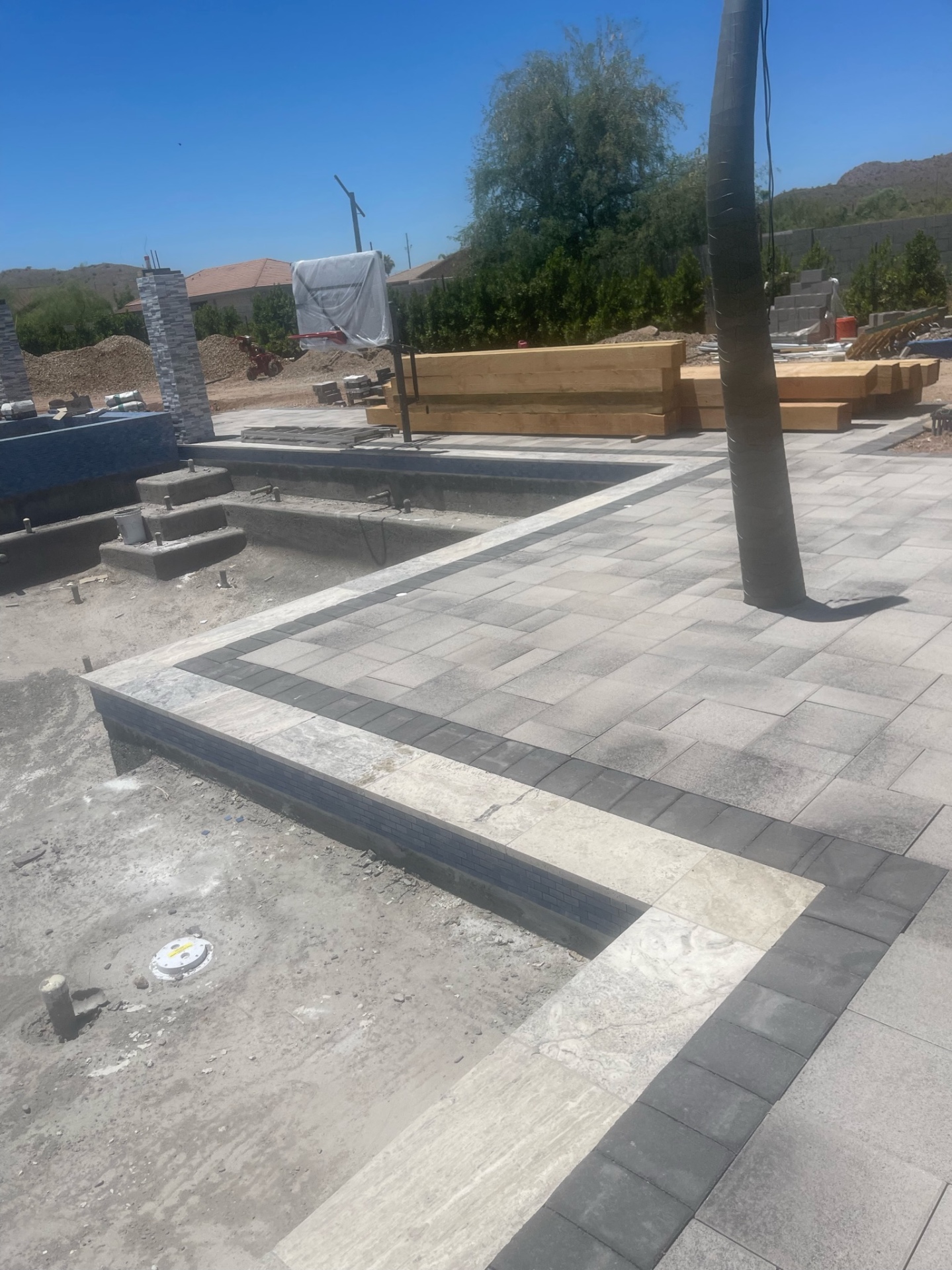 Close-up of large-format gray pavers with travertine and dark band borders, pergola lumber stacked behind