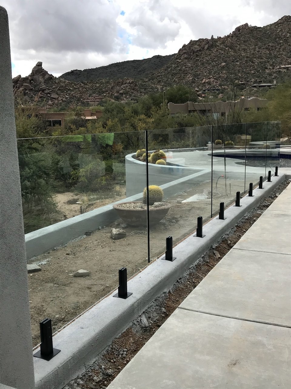 Long run of glass fence panels on black spigots along a painted concrete curb, framing a desert pool deck and granite mountain backdrop.