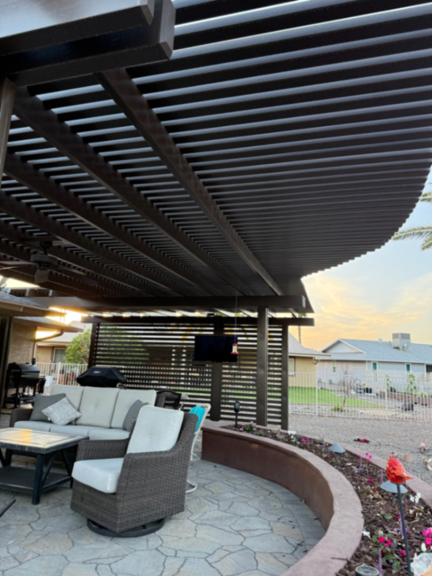 Thorn residence, Mesa AZ — louvered pergola over flagstone-pattern paver patio with a curved raised planter, mounted TV, and wood slat privacy wall.