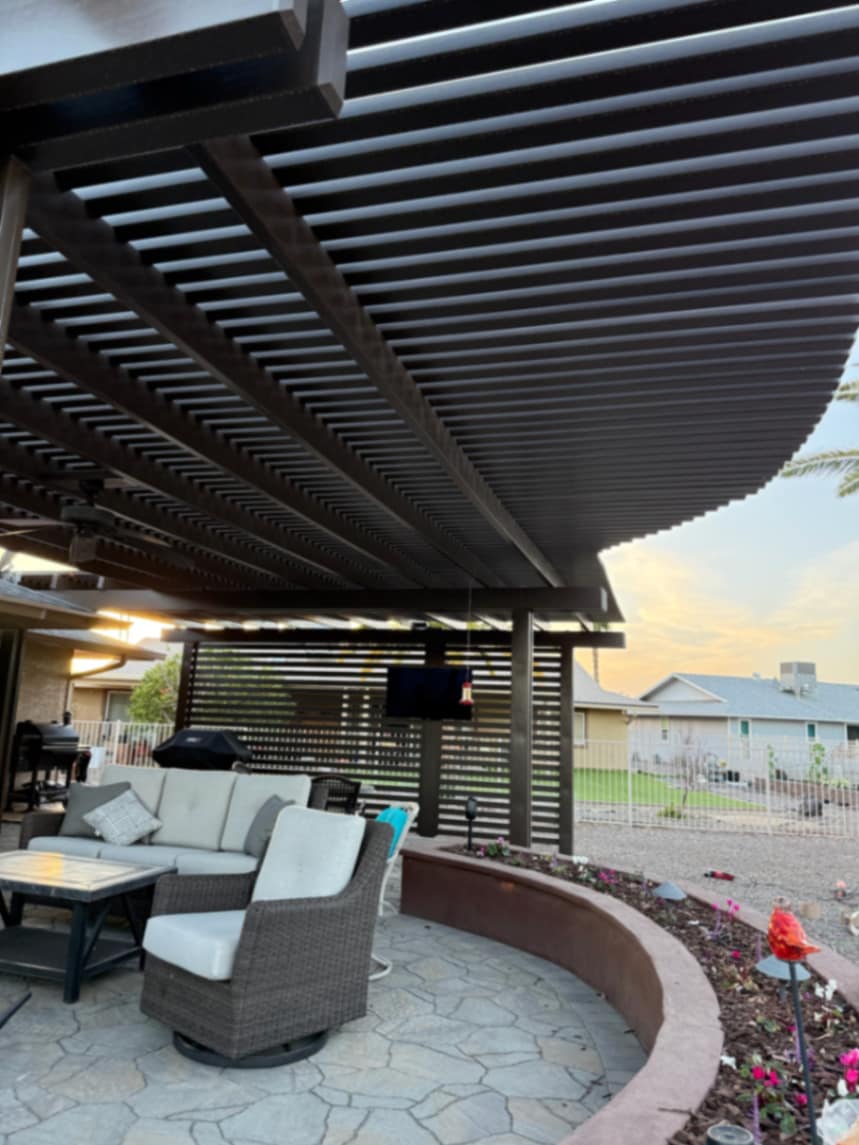 Thorn residence Mesa AZ louvered pergola over flagstone-pattern paver patio with curved raised stucco planter, mounted TV, and wood slat privacy wall at dusk