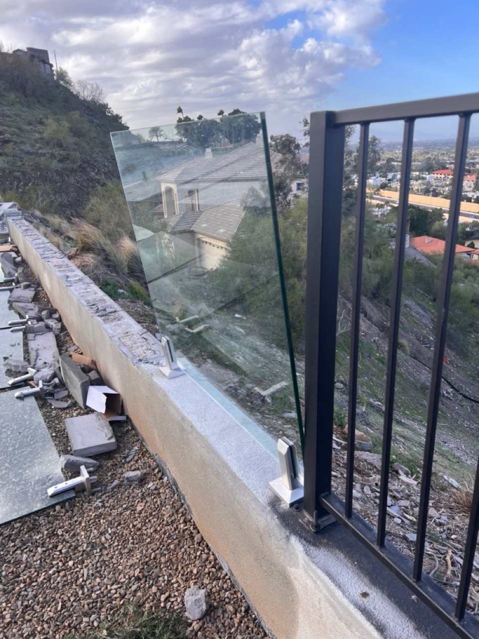 A single glass panel still leaning against a wrought-iron gate with adjacent bolt-down spigots on a crumbling stucco wall — same install as the failed panels.
