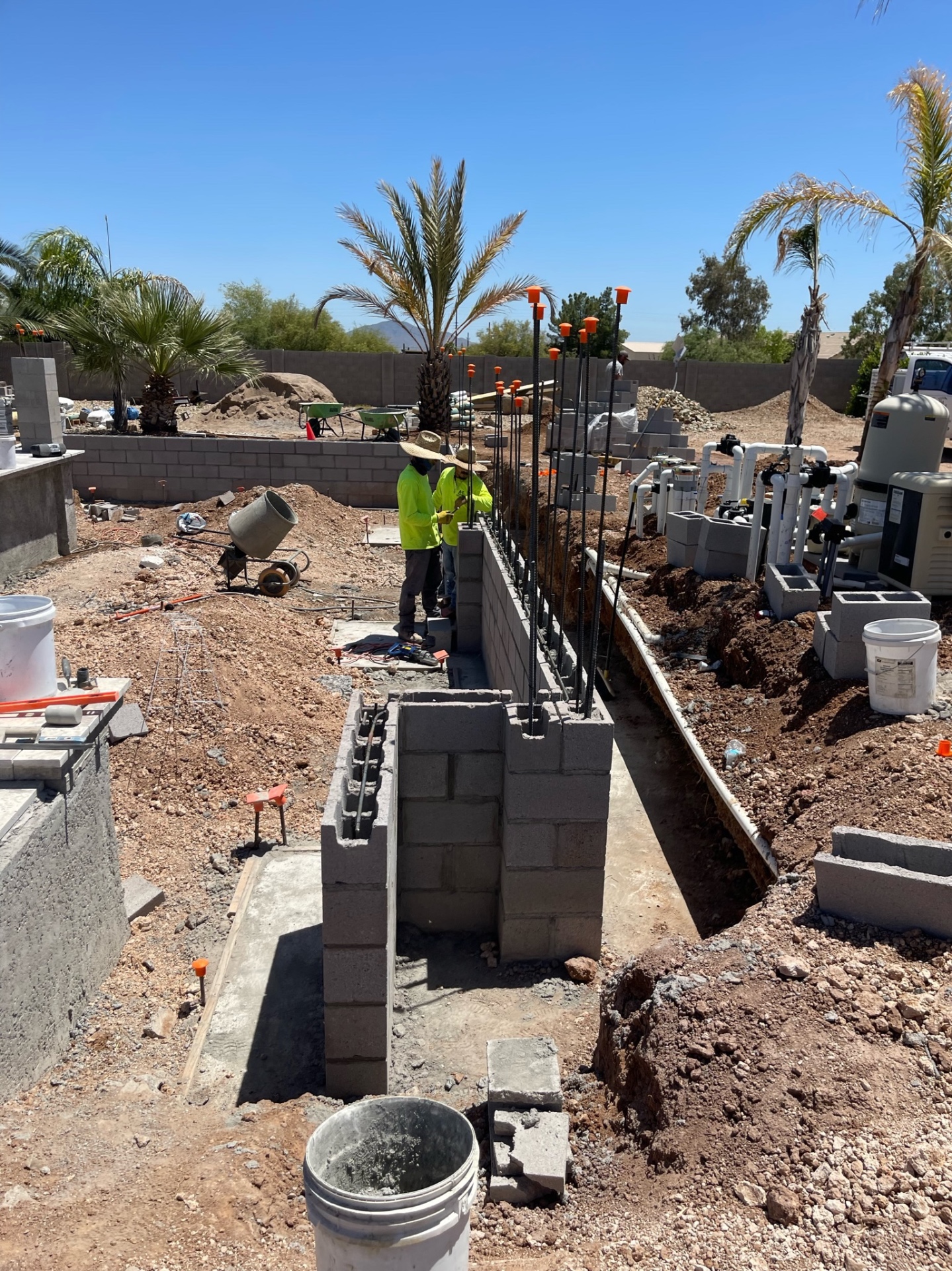 Two crew members setting block on a long retaining wall with pool equipment and trenching on the right side of the yard