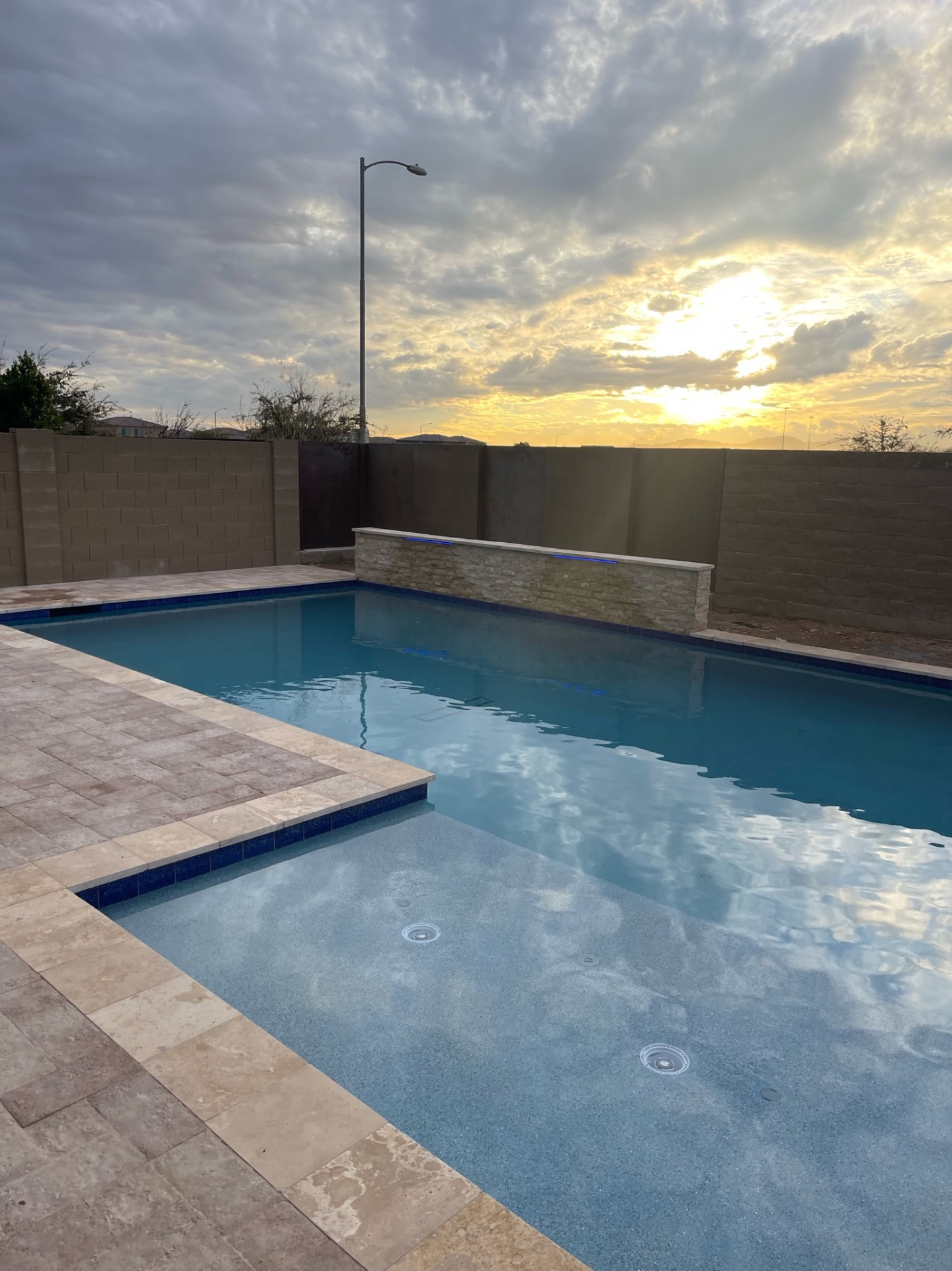 Peoria rectangular pool with pebble finish, stacked-stone raised sheer-descent water wall, blue-glass waterline tile and travertine paver deck at sunset