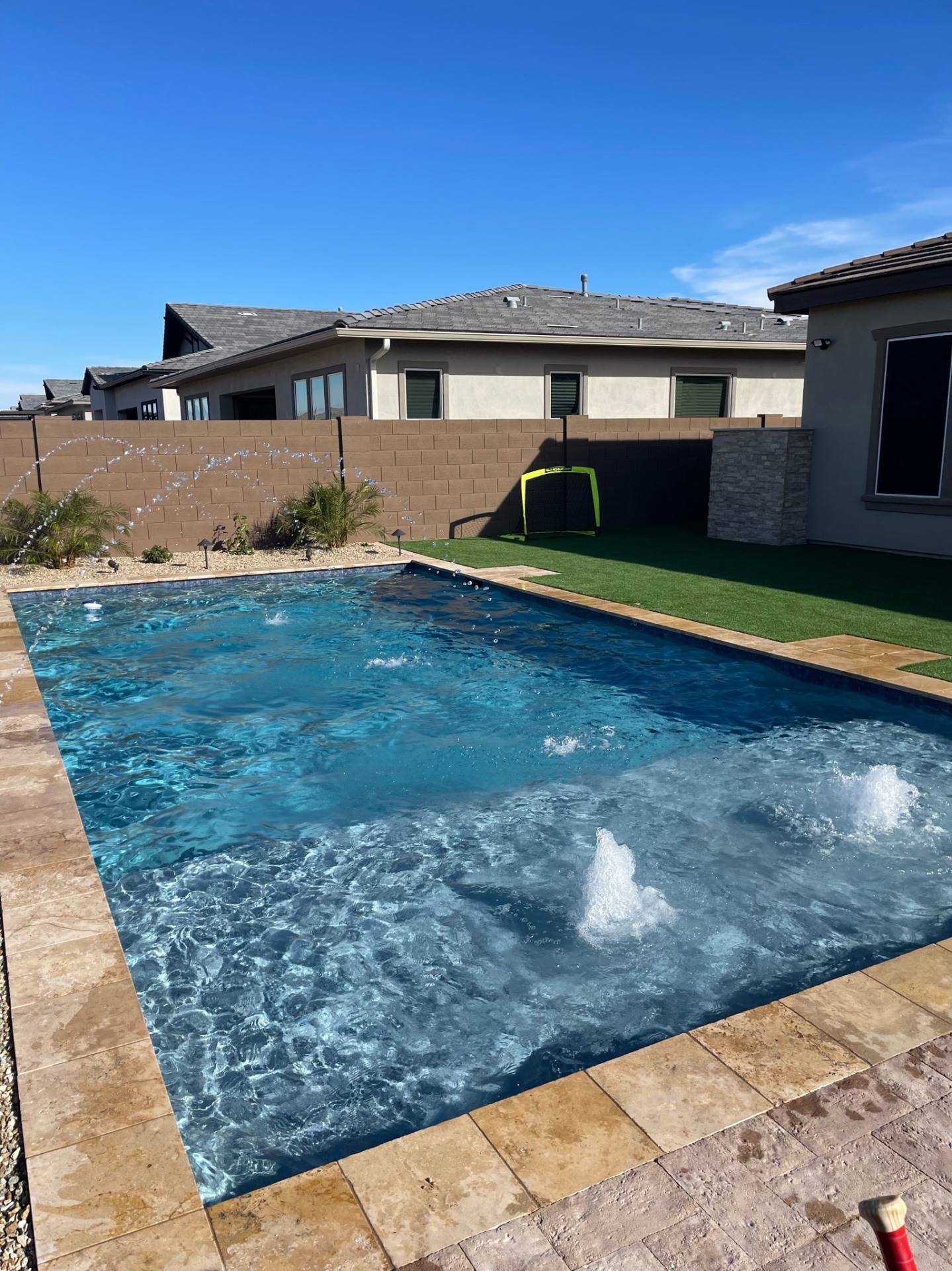 Peoria rectangular pool with deck-jet water arcs, in-pool bubblers, travertine paver coping and artificial turf side lawn against block wall by AE Outdoor Living