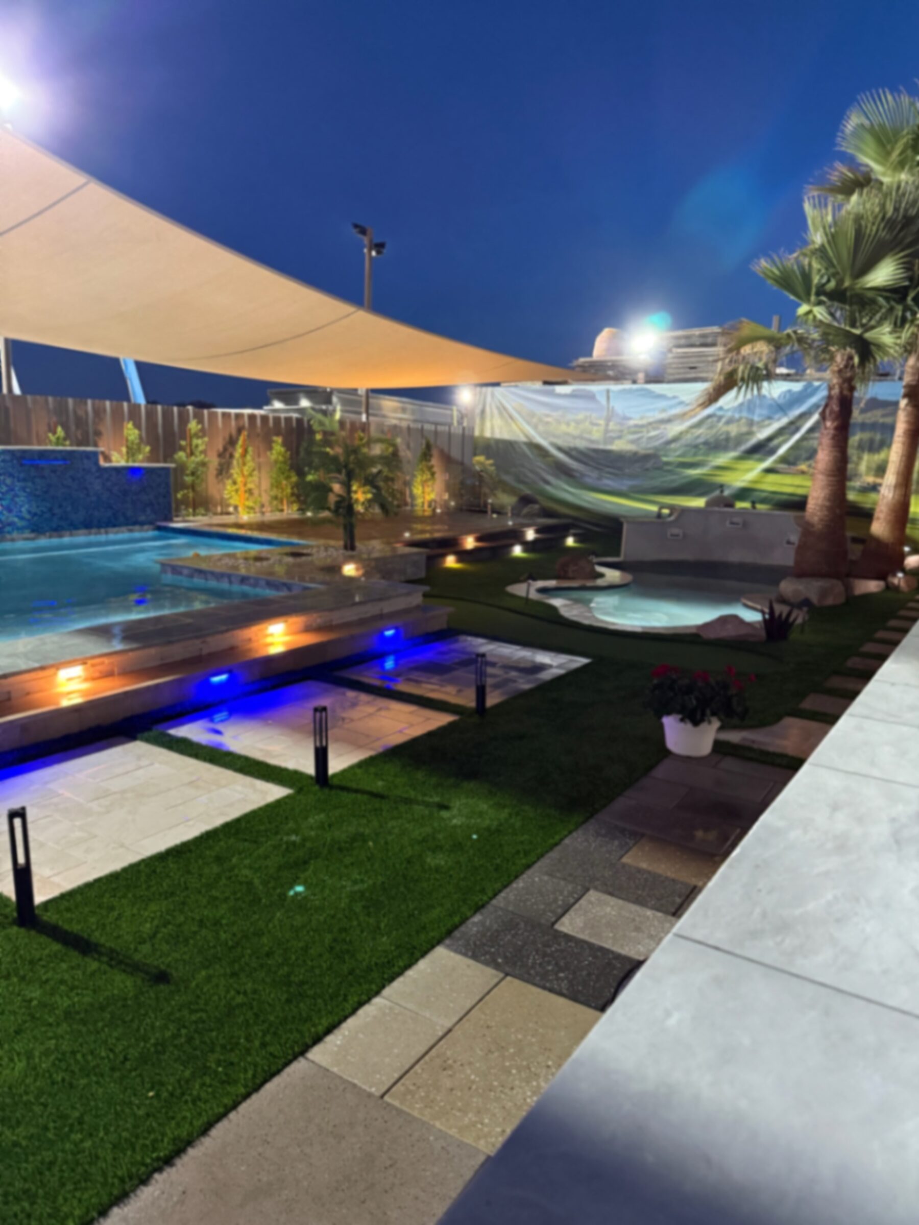 Wide dusk view of display pool with glass-tile spillover wall, paver-and-turf decking, attached spa, and palm trees under shade sails