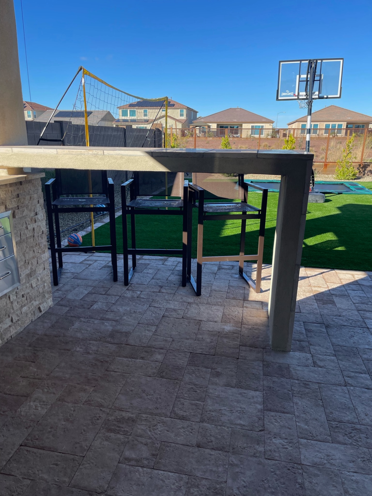 Peoria paver patio bar-height seating area with stacked-stone bar island, travertine bar top and artificial turf sport lawn beyond by AE Outdoor Living