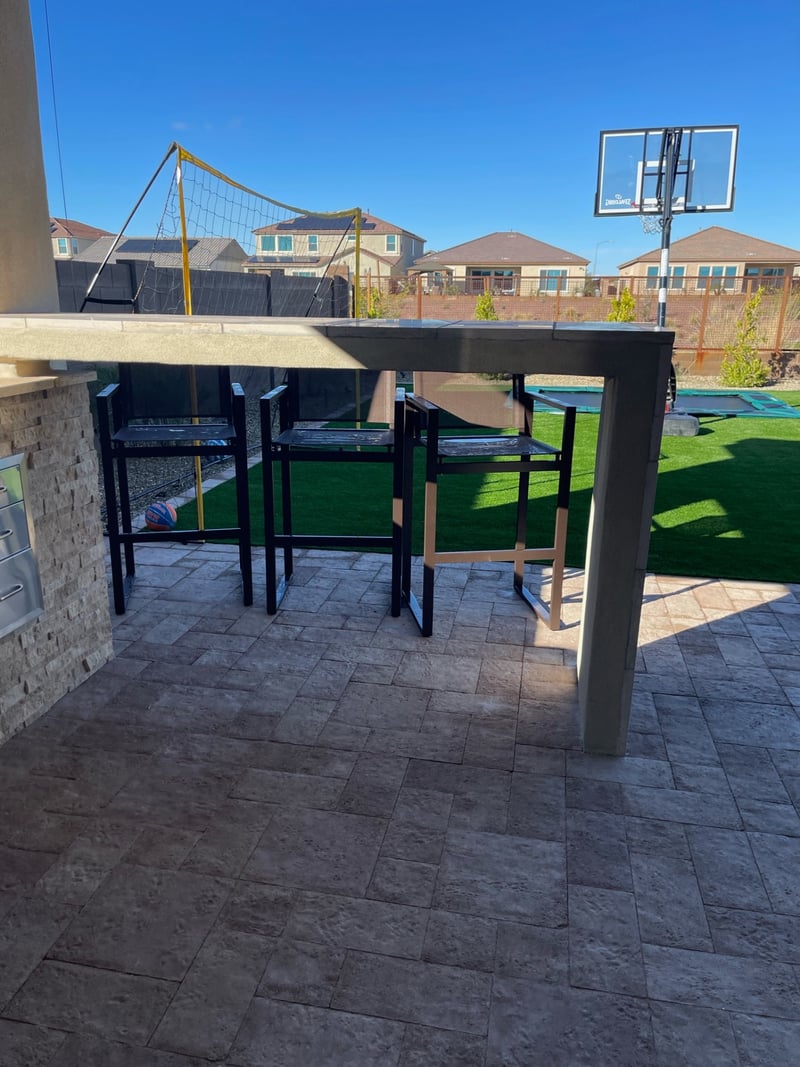 Peoria paver patio bar-height seating area with stacked-stone bar island, travertine bar top and artificial turf sport lawn beyond by AE Outdoor Living