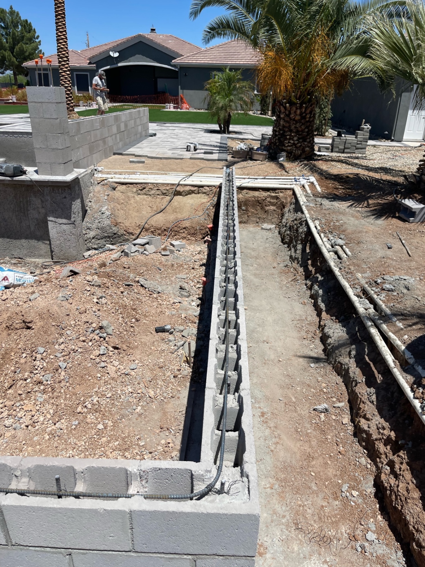 Deep block foundation trench with vertical rebar running along the side yard past palms toward the front house and paver walkway