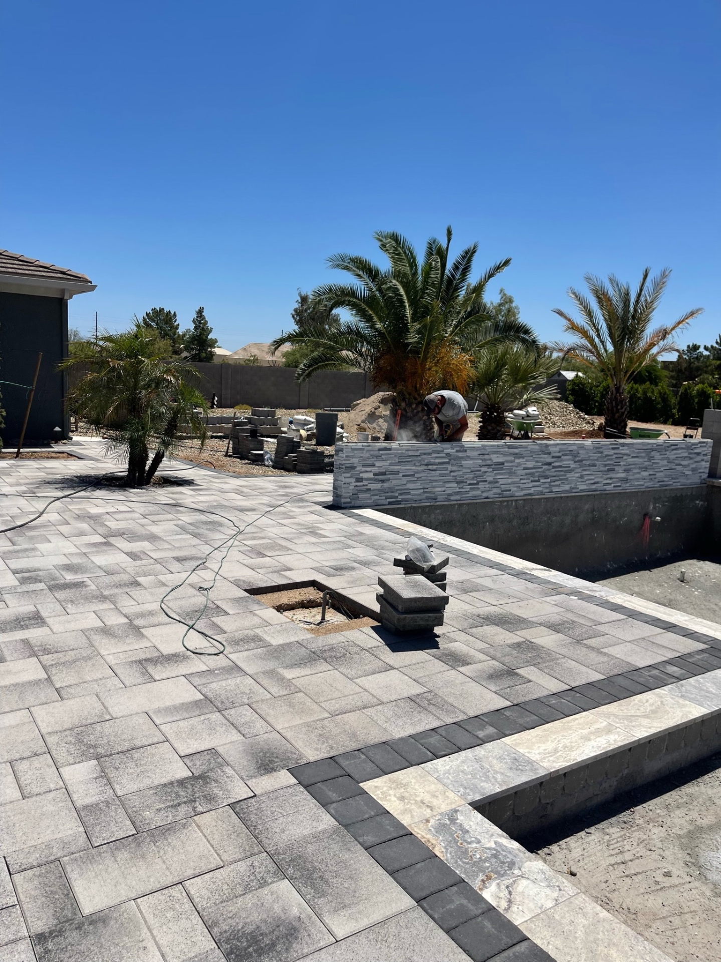 Large-format gray paver deck being installed around the pool with a crew member cutting stacked-stone veneer at the raised wall