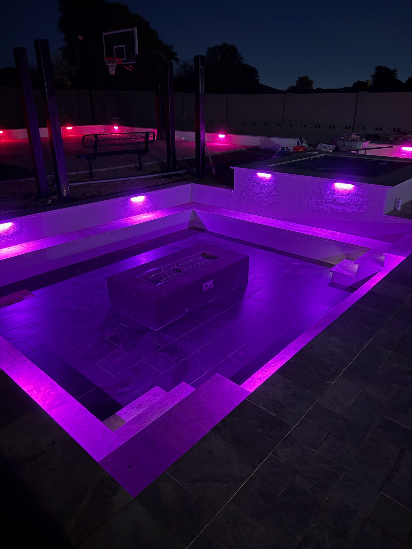 Sunken paver conversation lounge with linear fire pit, raised spa, and purple LED wall lights beside a sport court at night.