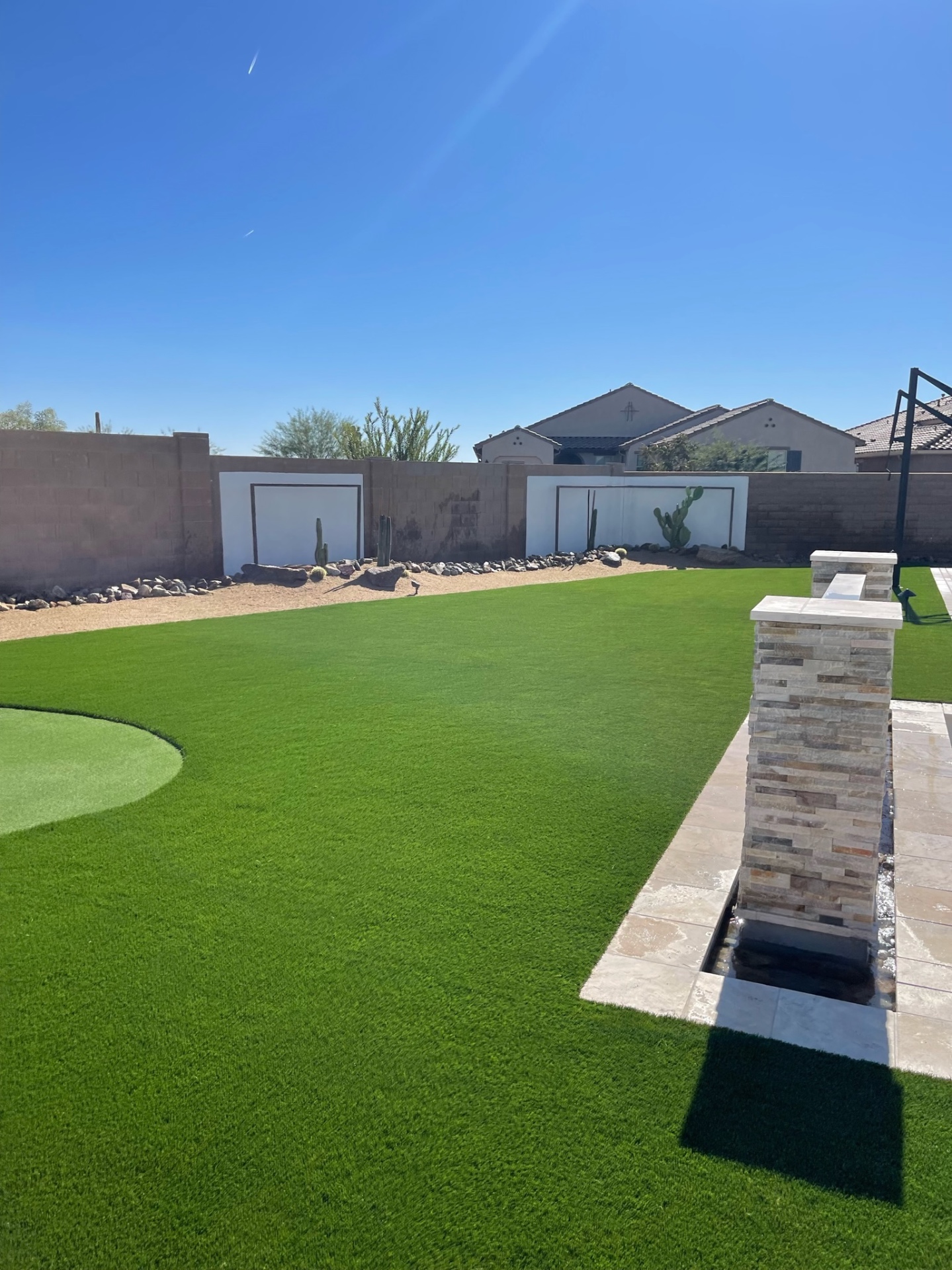Goodyear backyard artificial turf lawn with putting green and stacked-stone water feature column against desert rockwork border