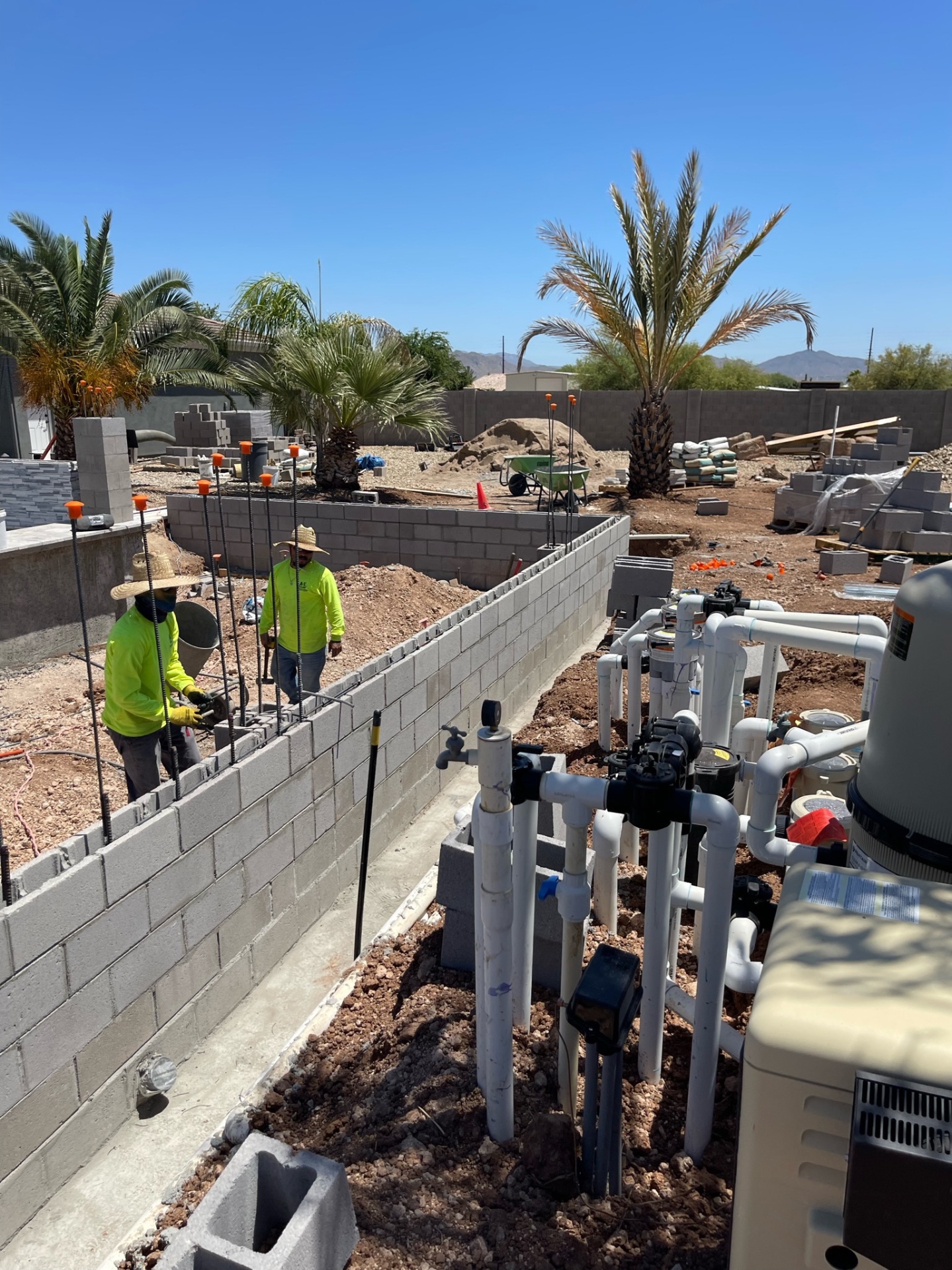 Two AE crew members in safety yellow laying CMU block on a perimeter wall with pool equipment manifold in foreground