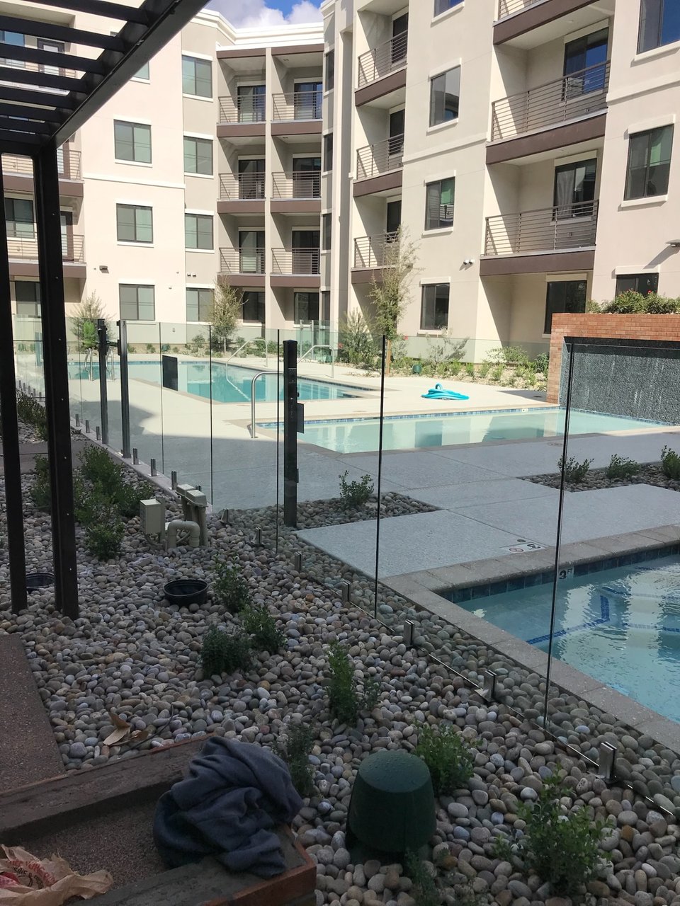 Glass fence panels running past a spa and pool, with a metal canopy structure and multifamily residences in the background.