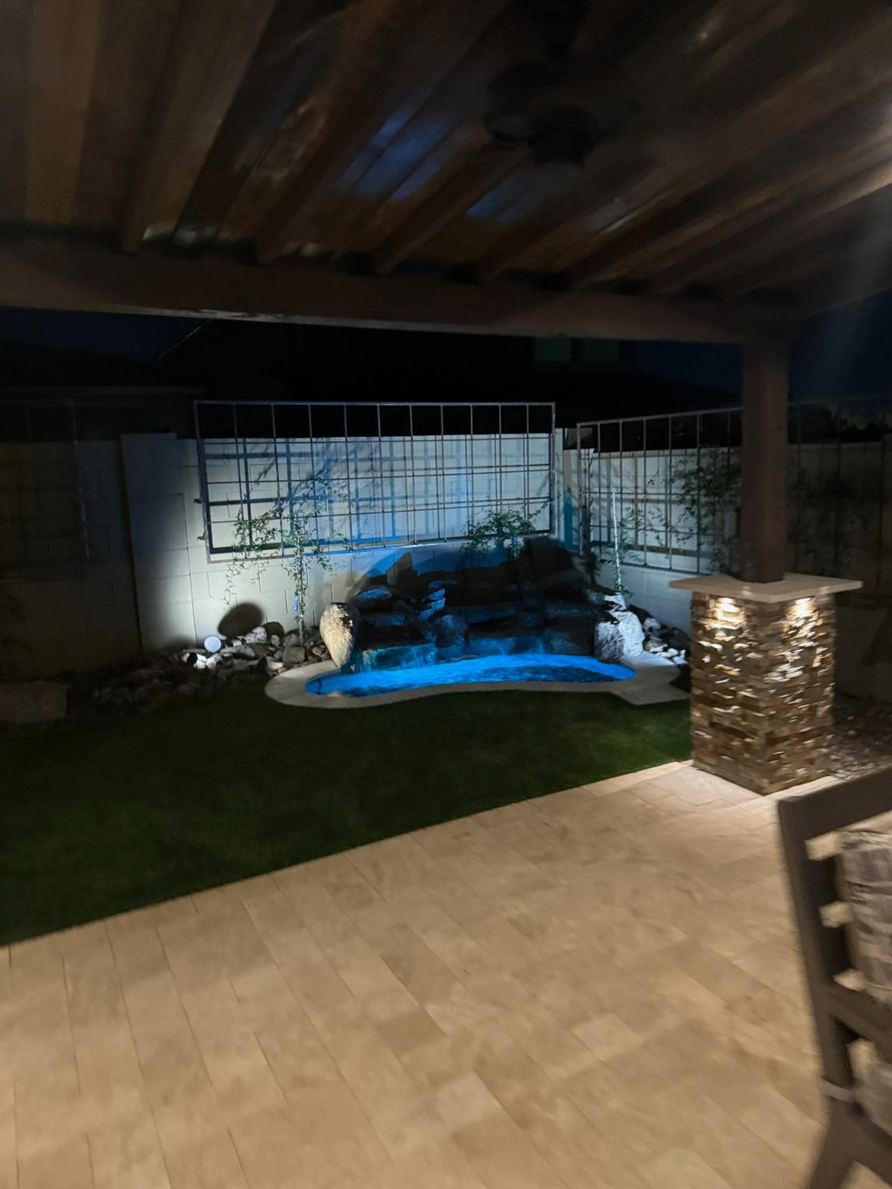 Small freeform spool with boulder waterfall and blue water light viewed from beneath a wood-lined covered patio over travertine pavers