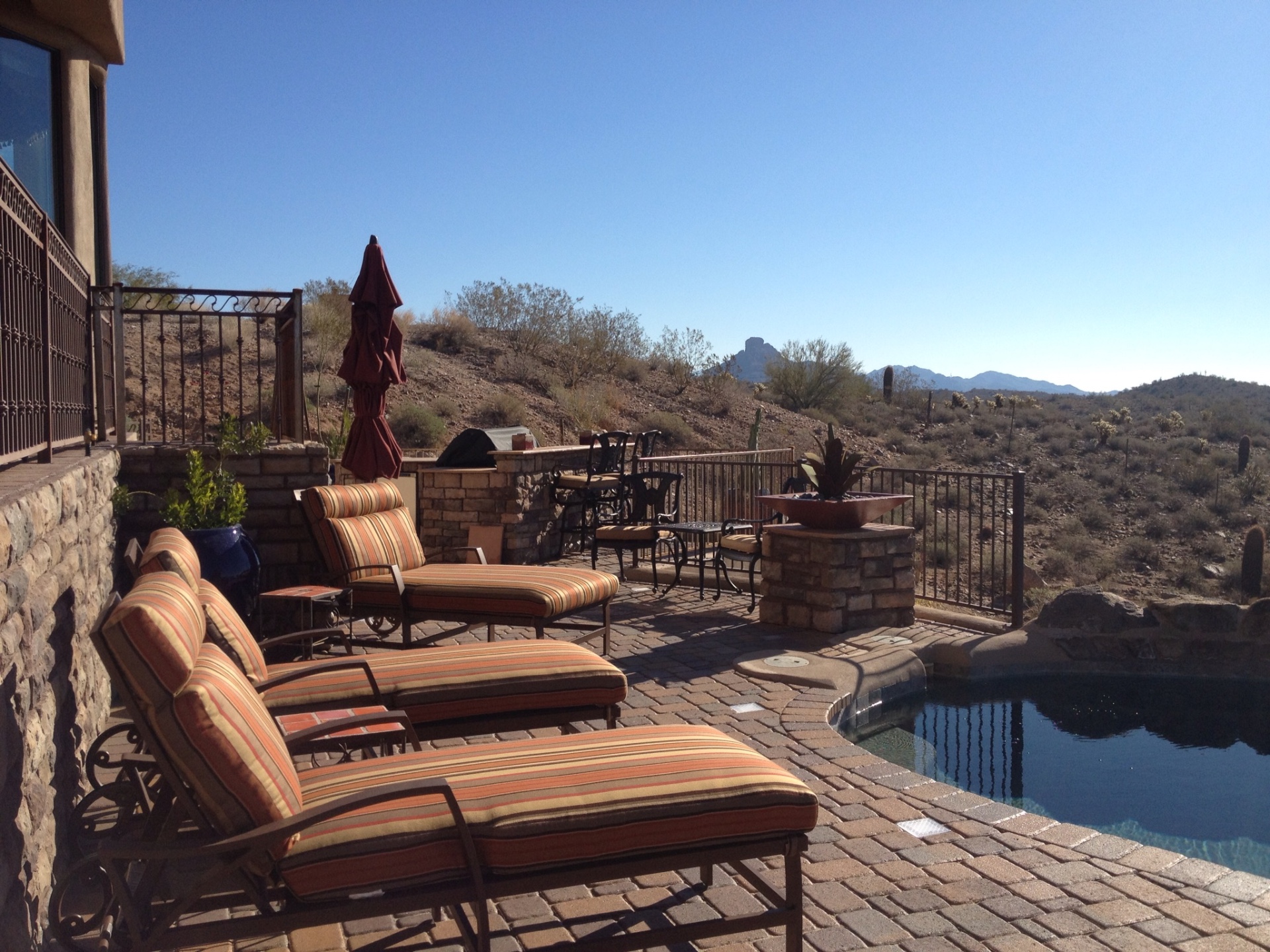 Fountain Hills upper paver terrace with stacked-stone bar-height BBQ island, wrought-iron view fencing and infinity pool below overlooking desert ridgeline