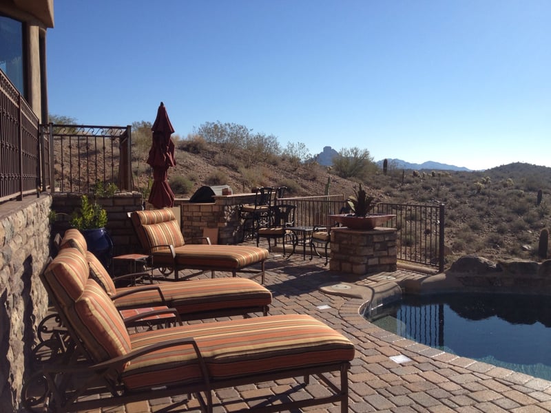 Fountain Hills upper paver terrace with stacked-stone bar-height BBQ island, wrought-iron view fencing and infinity pool below overlooking desert ridgeline