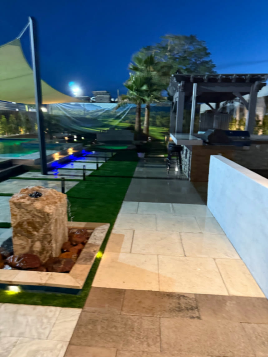 Walkway through the display yard with fountain, sail shade, turf strips, and outdoor kitchen lit for evening viewing