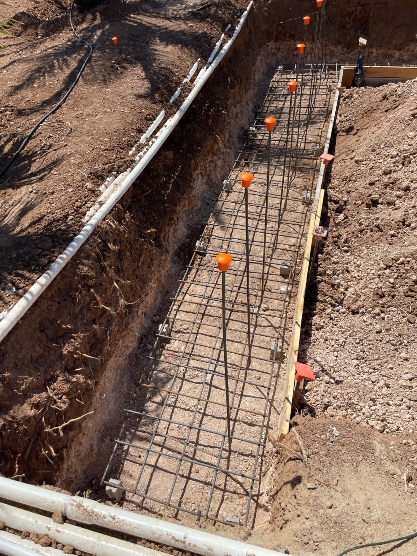 Close view down a narrow footing trench showing tied steel, spacers, and orange safety caps along the top bars