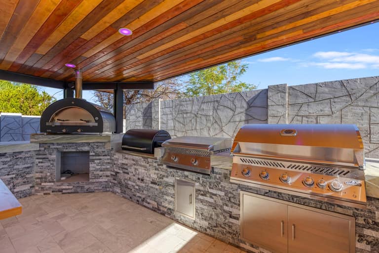 Outdoor cooking line with a pizza oven and multiple built-in appliances beneath a covered pavilion.