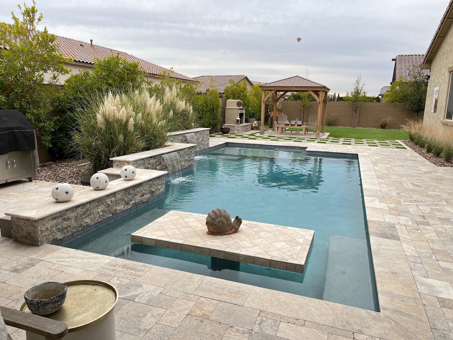 Vask residence, The Meadows, Peoria AZ — pool corner with travertine tanning-ledge platform, stacked-stone raised wall with spillway, and cedar gazebo with stucco fireplace beyond.