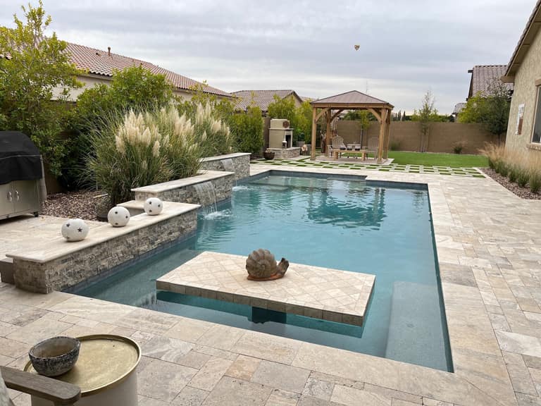 Vask residence The Meadows Peoria AZ pool corner with travertine tanning-ledge platform, stacked-stone raised wall with spillway, and cedar gazebo with stucco fireplace beyond