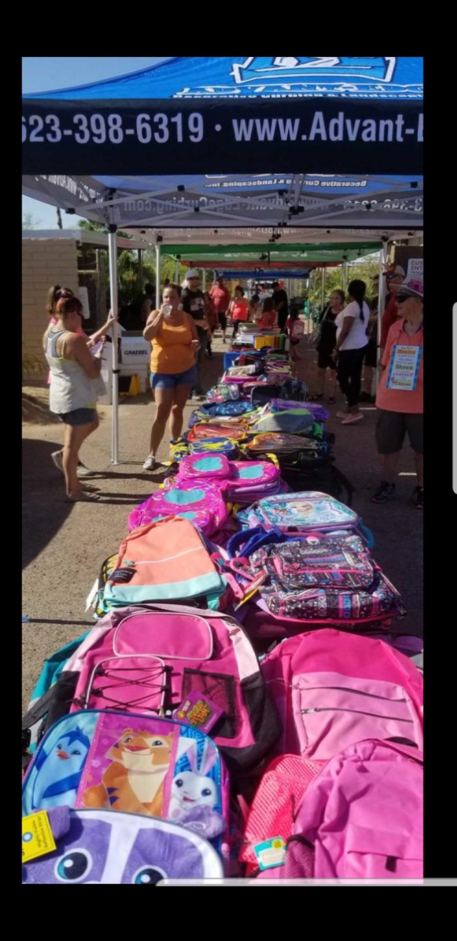 Long table of colorful kids' backpacks under partner tents at an outdoor distribution event in Arizona