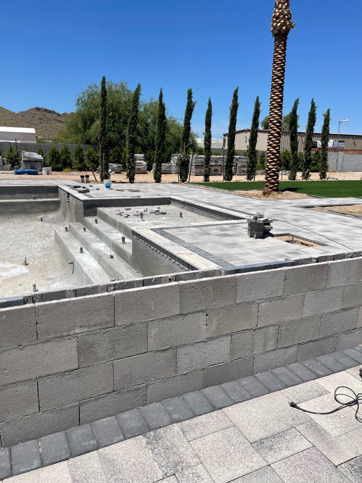 Pool shell with paver coping started, raised tiled spa, and stacked pavers staged beside palms ready for install