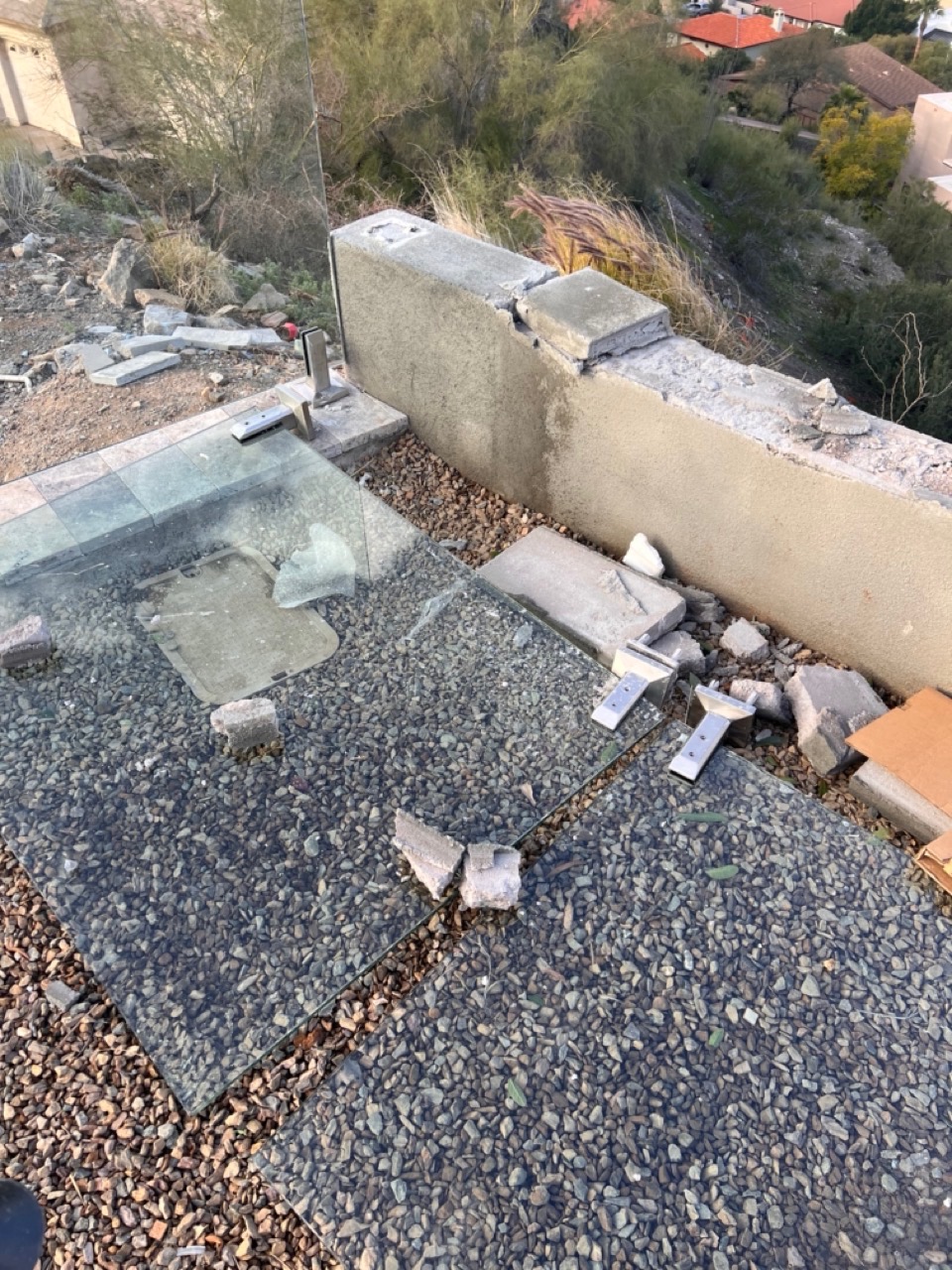 A tempered glass pool fence panel lying flat in landscape gravel with the bolt-down spigot brackets still attached and chunks of the wall cap that pulled out with it.