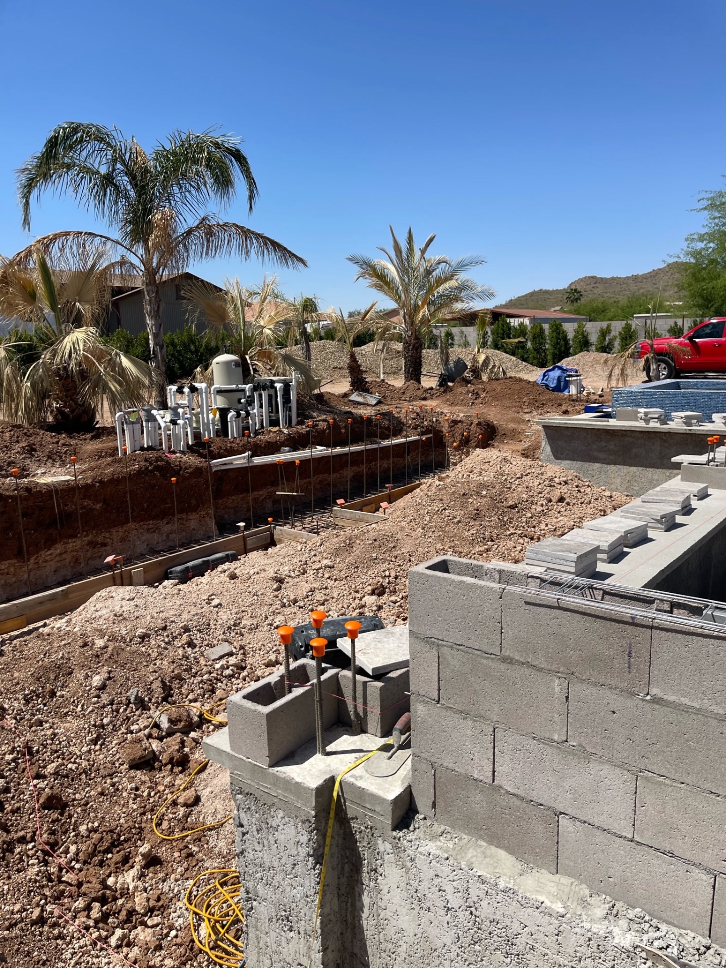 Wide jobsite view showing open shell work, block walls, equipment plumbing cluster, soil stockpiles, and a red AE truck at the far edge