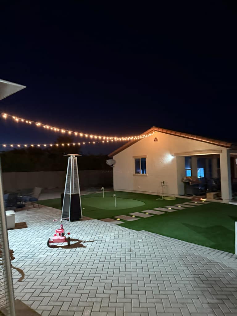 Herringbone paver patio meeting artificial-turf putting green with paver steppers and warm string lights overhead.