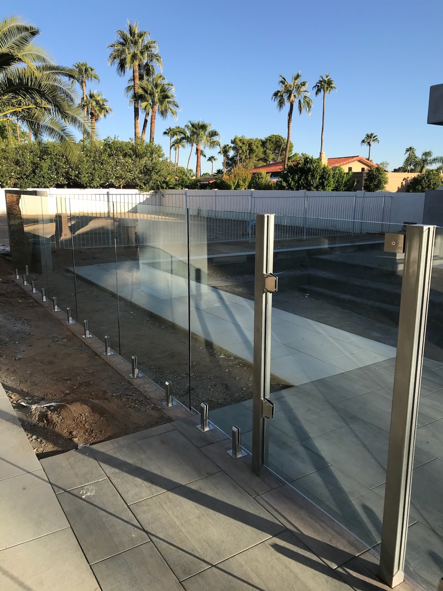 Frameless glass pool fence and stainless gate framing a new pool deck — code-height tempered panels on spigot mounts with palms and white perimeter fence beyond.