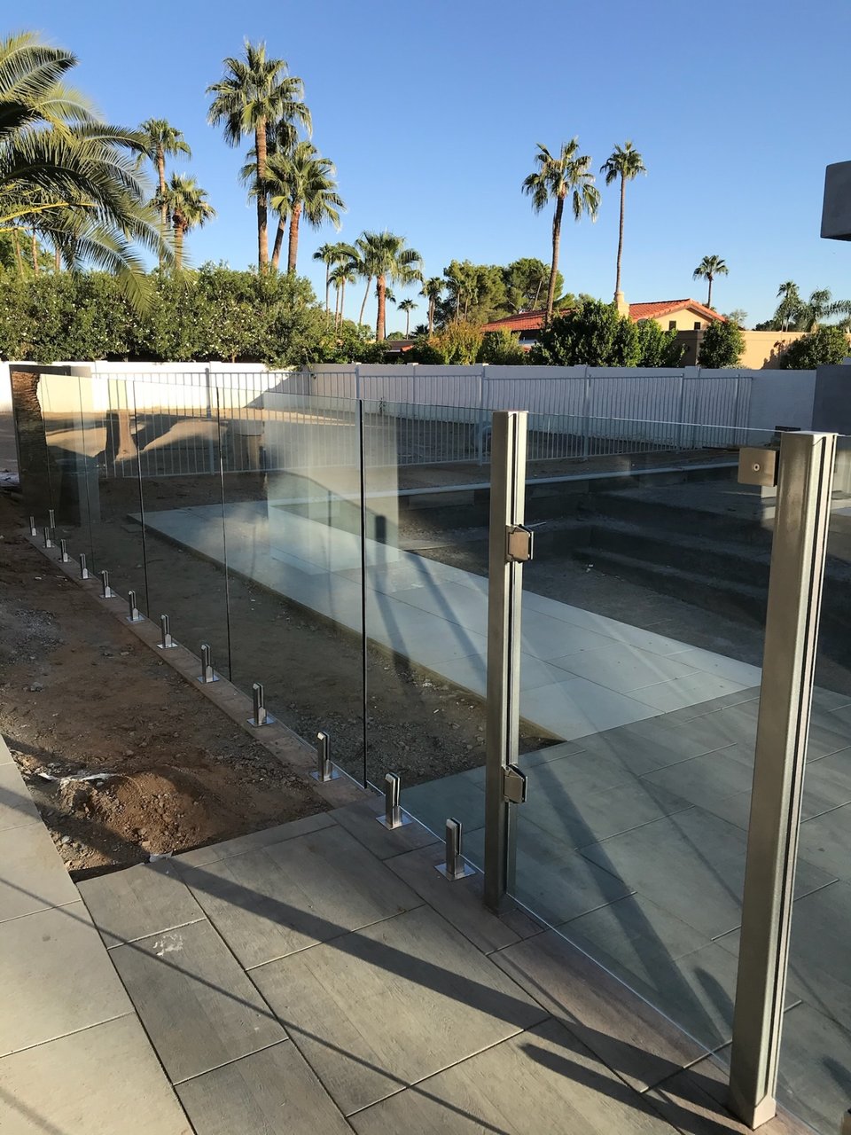 Frameless glass fencing and a metal-framed gate along a pool deck, with palms and a white perimeter fence beyond.