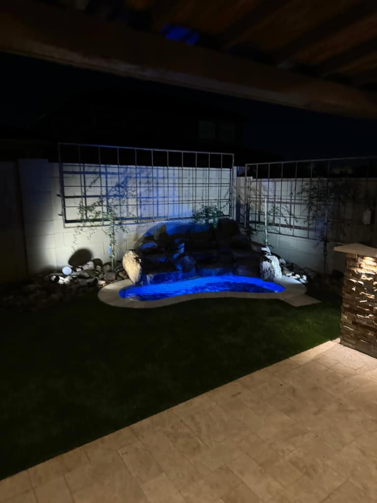 Closer night view of the spool and rock waterfall framed by metal trellis panels, climbing vines, turf, and travertine paving