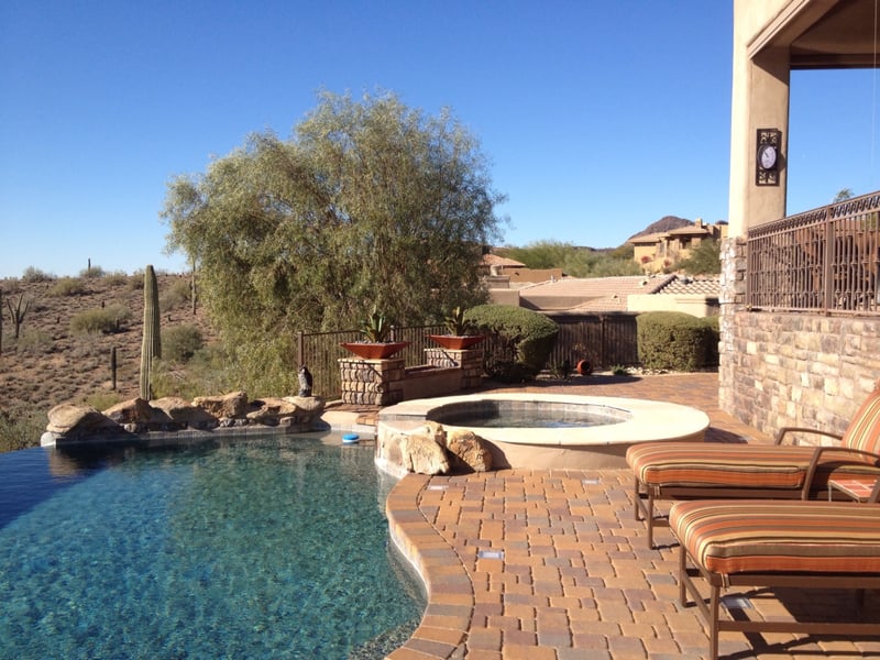 Fountain Hills negative-edge infinity pool with raised travertine spa, stacked-stone waterfall boulder wall and paver pool deck overlooking McDowell Mountain desert by AE Outdoor Living